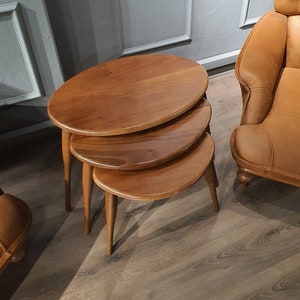 May include: Three wooden nesting tables with a rounded oval top. The tables are stacked on top of each other and have a light brown finish.