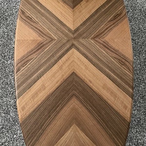 May include: Oval wooden coffee table with a chevron pattern. The tabletop features alternating light and dark brown wood planks arranged in a symmetrical design. The table is set against a gray carpet.