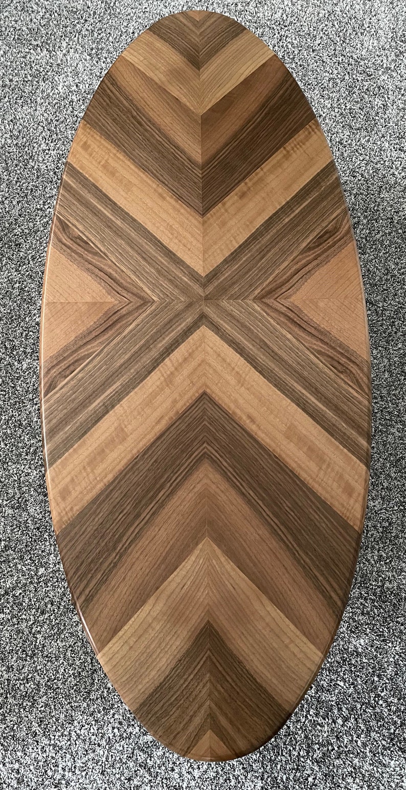 May include: Oval wooden coffee table with a chevron pattern. The tabletop features alternating light and dark brown wood planks arranged in a symmetrical design. The table is set against a gray carpet.