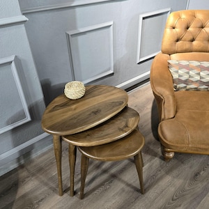 May include: Three wooden nesting tables with a rounded, oval shape. The tables are made of dark brown wood and have a natural finish. The tables are stacked on top of each other, with the largest table on the bottom and the smallest table on top.