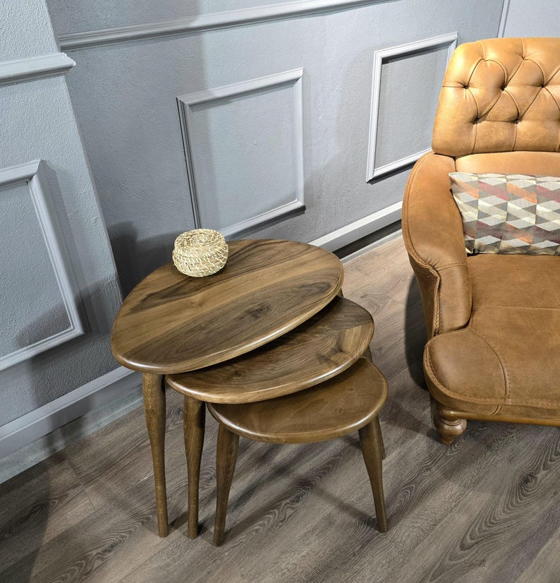 May include: Three wooden nesting tables with a rounded, oval shape. The tables are made of dark brown wood and have a natural finish. The tables are stacked on top of each other, with the largest table on the bottom and the smallest table on top.