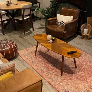 May include: A living room scene featuring a mid-century modern coffee table with a wooden chevron pattern, set on a patterned rug. A brown leather armchair with a decorative pillow sits nearby. The room includes a round wooden table and a leather ottoman.