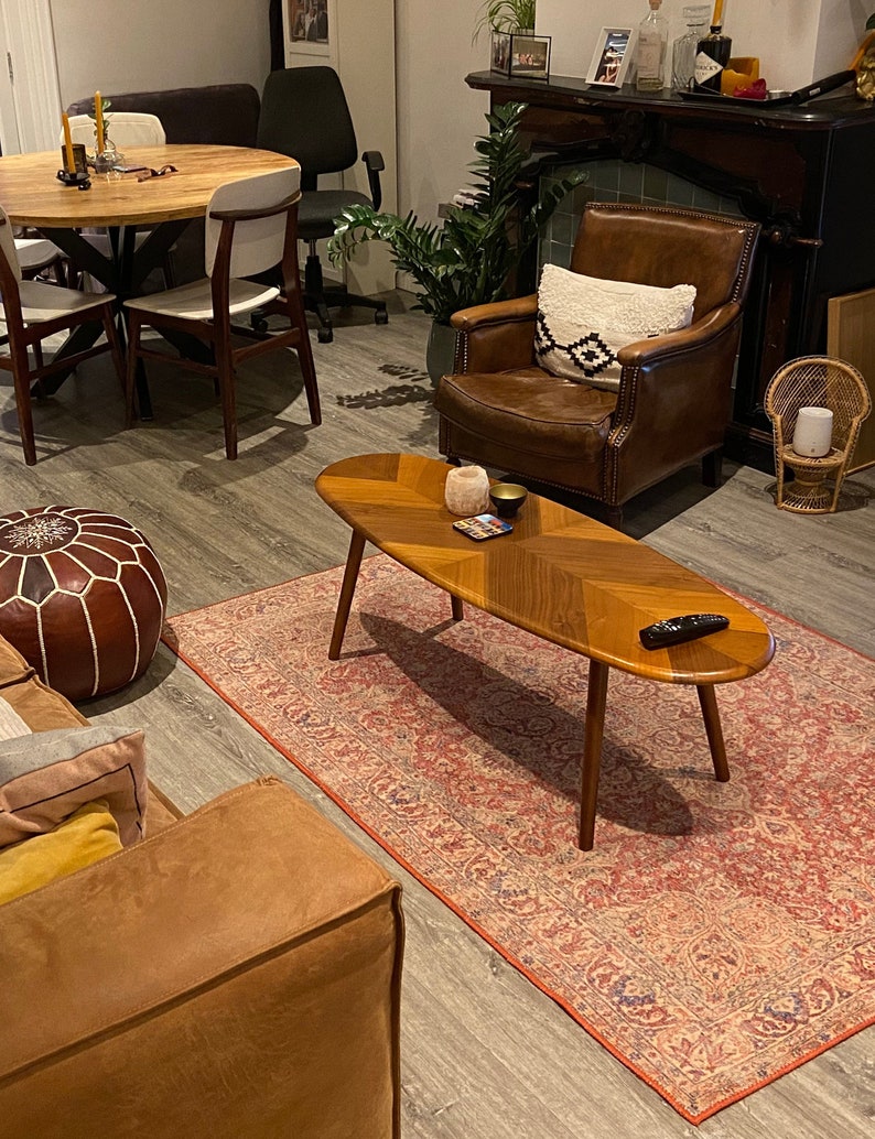 May include: A living room scene featuring a mid-century modern coffee table with a wooden chevron pattern, set on a patterned rug. A brown leather armchair with a decorative pillow sits nearby. The room includes a round wooden table and a leather ottoman.