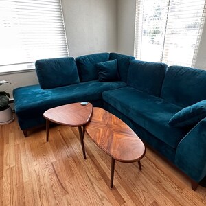 May include: A teal sectional sofa with a matching throw pillow and two wooden coffee tables. The coffee tables have a mid-century modern design with dark wood and angled legs. A potted plant sits near a window.