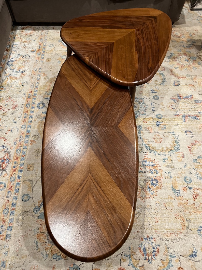 May include: Two-piece nesting coffee table set. The tables are made of wood with a dark brown finish and a chevron pattern. The top table is 24 inches long, and the bottom table is 36 inches long. The tables have a modern design.