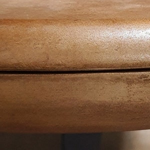 May include: Close-up of the edge of a round, wooden table top. The table top is a light brown color and has a smooth, polished finish.