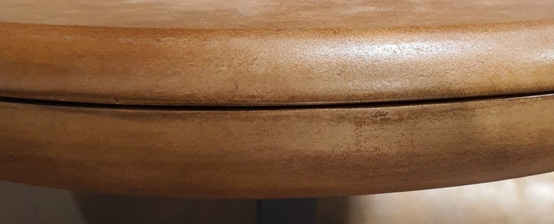 May include: Close-up of the edge of a round, wooden table top. The table top is a light brown color and has a smooth, polished finish.