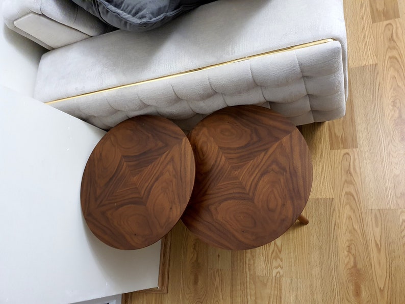 May include: Two round, dark brown wooden side tables with a unique geometric design. The tables are placed on a light-coloured wooden floor, near a light-coloured sofa with a gold trim.