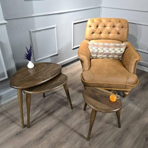 May include: Two wooden nesting tables with a rounded, leaf-shaped top. The tables are brown and have a natural wood finish. The tables are sitting on a light brown wood floor. A brown leather armchair with a tufted back and a patterned pillow is in the background.