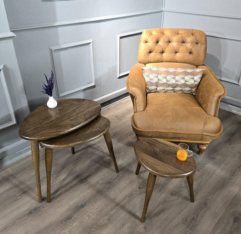 May include: Two wooden nesting tables with a rounded, leaf-shaped top. The tables are brown and have a natural wood finish. The tables are sitting on a light brown wood floor. A brown leather armchair with a tufted back and a patterned pillow is in the background.