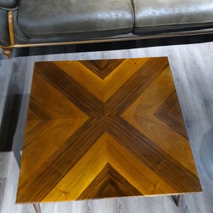 May include: Square wooden coffee table with a geometric pattern in shades of brown and tan. The tabletop features a symmetrical design with angled lines, creating a starburst effect. The table has four short, tapered legs.