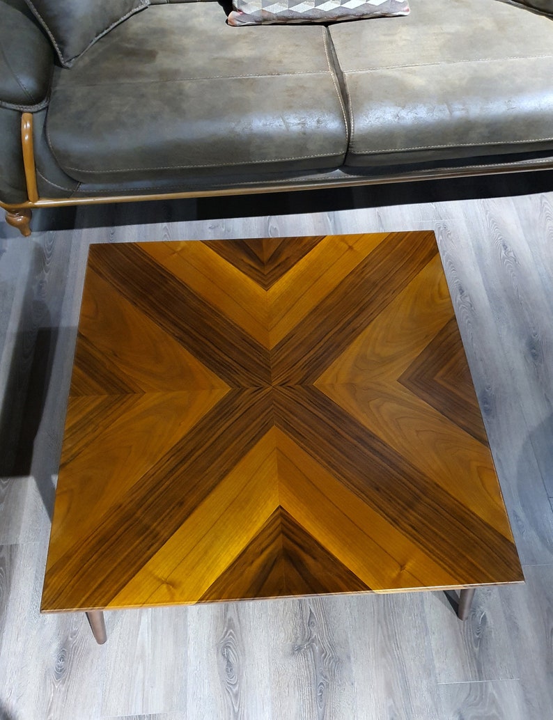 May include: Square wooden coffee table with a geometric pattern in shades of brown and tan. The tabletop features a symmetrical design with angled lines, creating a starburst effect. The table has four short, tapered legs.