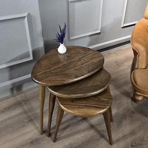 May include: Three wooden nesting tables with a rounded oval top. The tables are made of dark brown wood with a natural wood grain. The tables are arranged in a stack, with the largest table on the bottom and the smallest table on top.