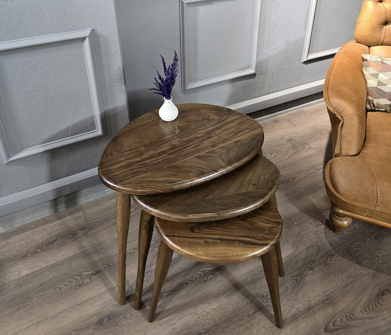 May include: Three wooden nesting tables with a rounded oval top. The tables are made of dark brown wood with a natural wood grain. The tables are arranged in a stack, with the largest table on the bottom and the smallest table on top.