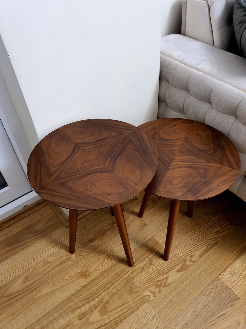 May include: Two round side tables with a dark brown, geometric patterned tabletop. Each table has three slender, tapered legs. The tables are placed on a light-coloured wooden floor.
