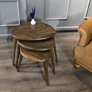 May include: A set of three wooden nesting tables with a rounded, oval-shaped top. The tables are made of dark brown wood and have a natural finish. The tables are stacked on top of each other, with the largest table on the bottom and the smallest table on top.