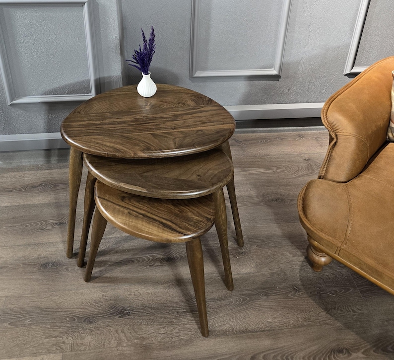 May include: A set of three wooden nesting tables with a rounded, oval-shaped top. The tables are made of dark brown wood and have a natural finish. The tables are stacked on top of each other, with the largest table on the bottom and the smallest table on top.