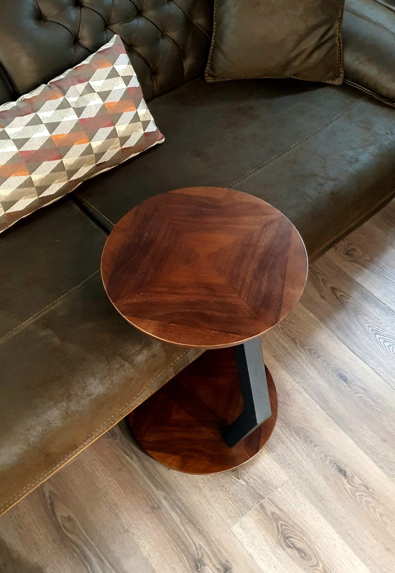 May include: A round wooden side table with a black metal base. The table top has a wood grain pattern and is dark brown in color. The table is sitting on a light brown wood floor.
