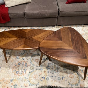 May include: Two wooden coffee tables with a unique design. One is oval, the other is triangular, both with a chevron pattern on the surface. The tables have tapered legs and are set on a patterned rug.