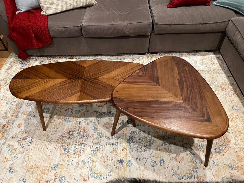 May include: Two wooden coffee tables with a unique design. One is oval, the other is triangular, both with a chevron pattern on the surface. The tables have tapered legs and are set on a patterned rug.