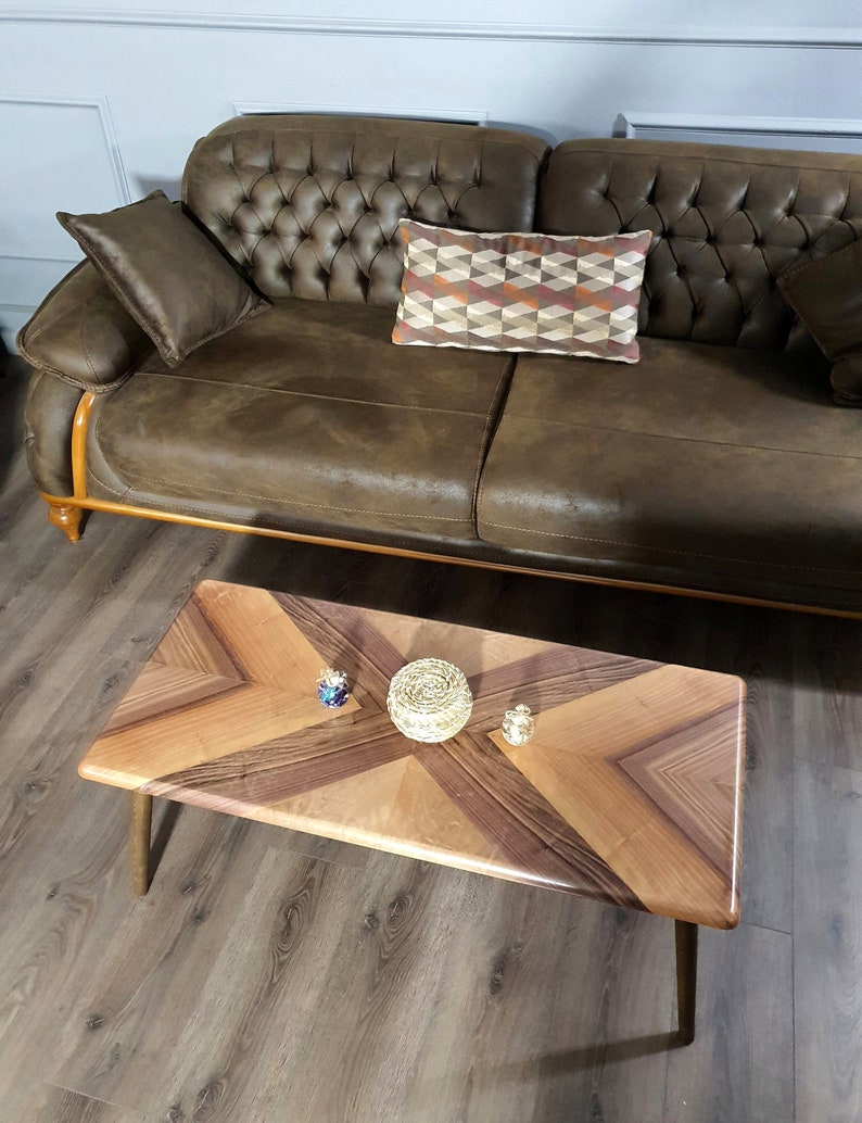 May include: A brown leather sofa with a tufted back and wooden trim. A rectangular pillow with a geometric pattern rests on the sofa. A wooden coffee table with a chevron design and decorative objects sits in front of the sofa.