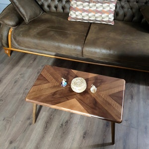 May include: A rectangular wooden coffee table with a chevron pattern in shades of brown. The table has four tapered legs and is accessorized with decorative items. A brown leather sofa and patterned pillow are in the background.