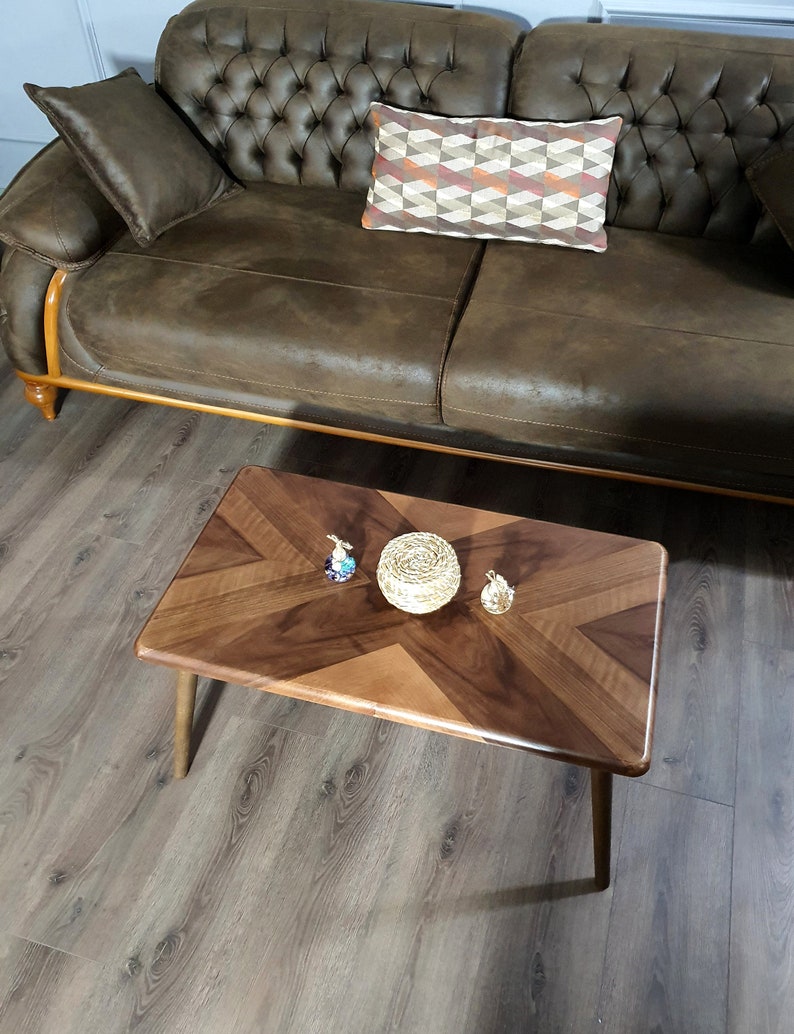 May include: A rectangular wooden coffee table with a chevron pattern in shades of brown. The table has four tapered legs and is accessorized with decorative items. A brown leather sofa and patterned pillow are in the background.