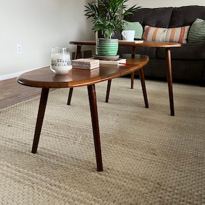 May include: Two mid-century modern style wooden coffee tables with dark brown legs and a light brown oval top. A green potted plant sits on the top table. A white candle and small decorative boxes are on the lower table. The tables are on a woven rug.