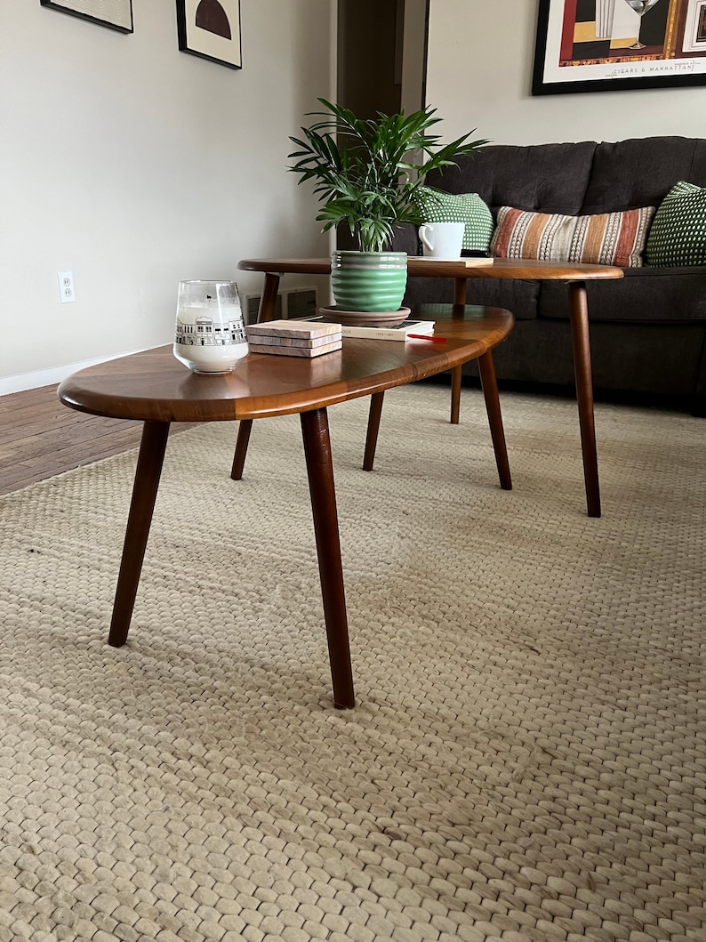 May include: Two mid-century modern style wooden coffee tables with dark brown legs and a light brown oval top. A green potted plant sits on the top table. A white candle and small decorative boxes are on the lower table. The tables are on a woven rug.