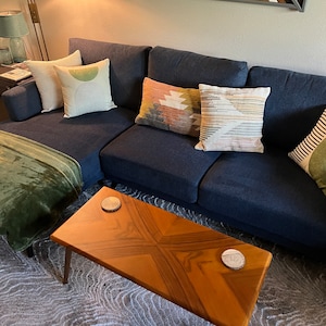 May include: A navy blue sectional sofa with several decorative throw pillows in various colors and patterns. A wooden coffee table with a starburst design sits in front of the sofa. Two coasters are on the table. A green blanket is draped over the sofa.