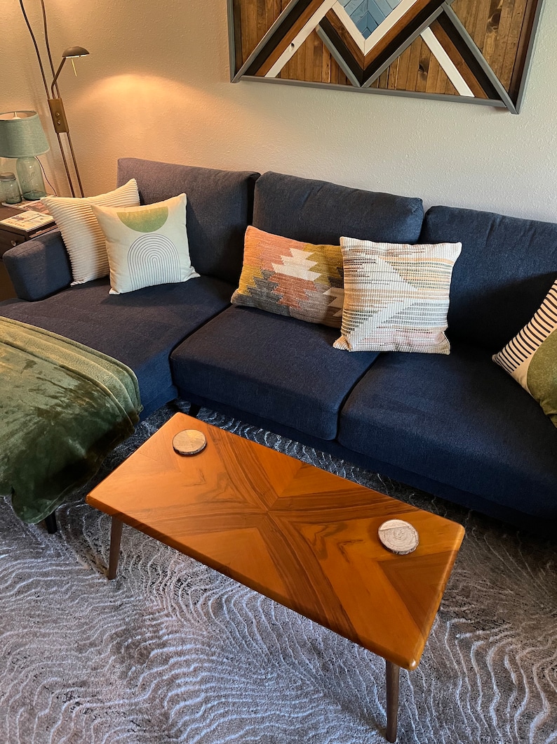 May include: A navy blue sectional sofa with several decorative throw pillows in various colors and patterns. A wooden coffee table with a starburst design sits in front of the sofa. Two coasters are on the table. A green blanket is draped over the sofa.
