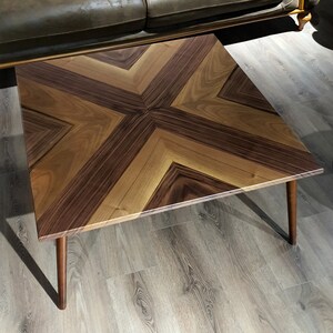 May include: A square coffee table with a geometric pattern of light and dark wood. The table has a modern design and is supported by four legs.
