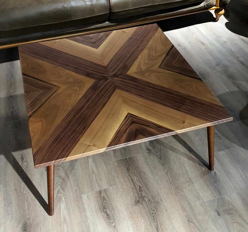 May include: A square coffee table with a geometric pattern of light and dark wood. The table has a modern design and is supported by four legs.