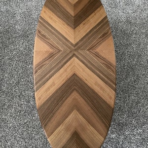 May include: Oval wooden coffee table with a chevron pattern in shades of brown. The table top is made of different types of wood, creating a unique and interesting design.