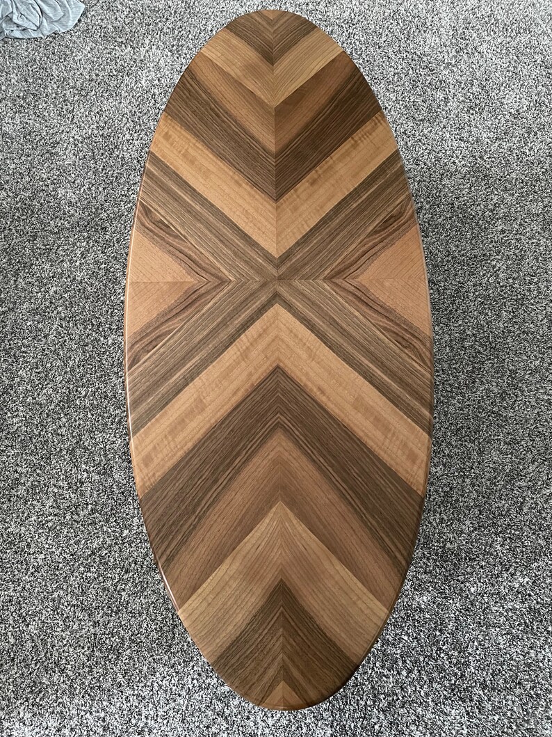May include: Oval wooden coffee table with a chevron pattern in shades of brown. The table top is made of different types of wood, creating a unique and interesting design.