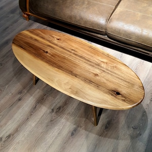 May include: Oval-shaped wooden coffee table with a natural wood finish and a light brown color. The table has a unique grain pattern and is supported by three thin, gold-colored legs.