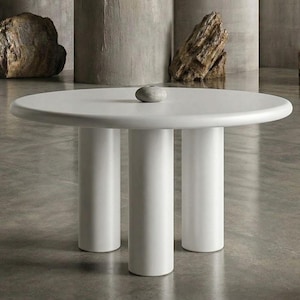 May include: A round white table with a smooth surface and three thick cylindrical legs. A small, smooth, grey stone object sits in the centre of the tabletop. The table features a clean, solid white finish throughout its entire structure.