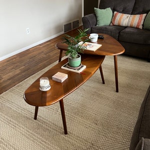 May include: Two mid-century modern wooden coffee tables. The top table is triangular, and the bottom table is oval. Both tables have dark brown legs and a light brown tabletop with a darker brown wood inlay. A small plant and a candle sit on the tables.