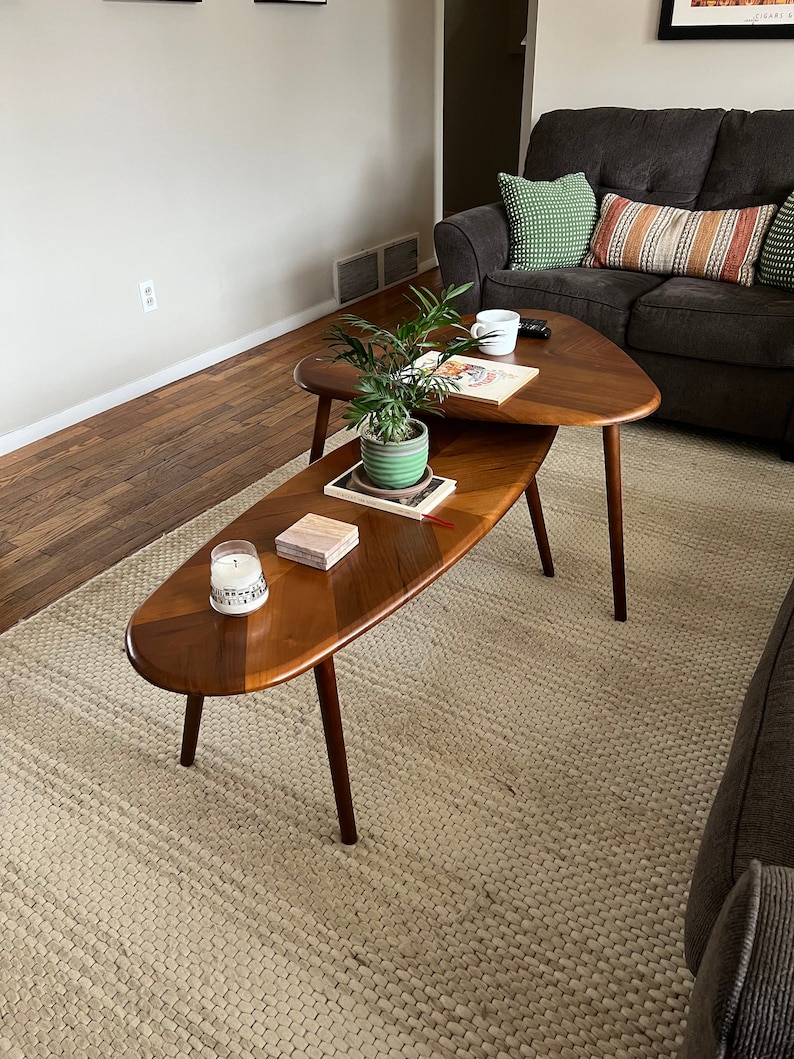 May include: Two mid-century modern wooden coffee tables. The top table is triangular, and the bottom table is oval. Both tables have dark brown legs and a light brown tabletop with a darker brown wood inlay. A small plant and a candle sit on the tables.