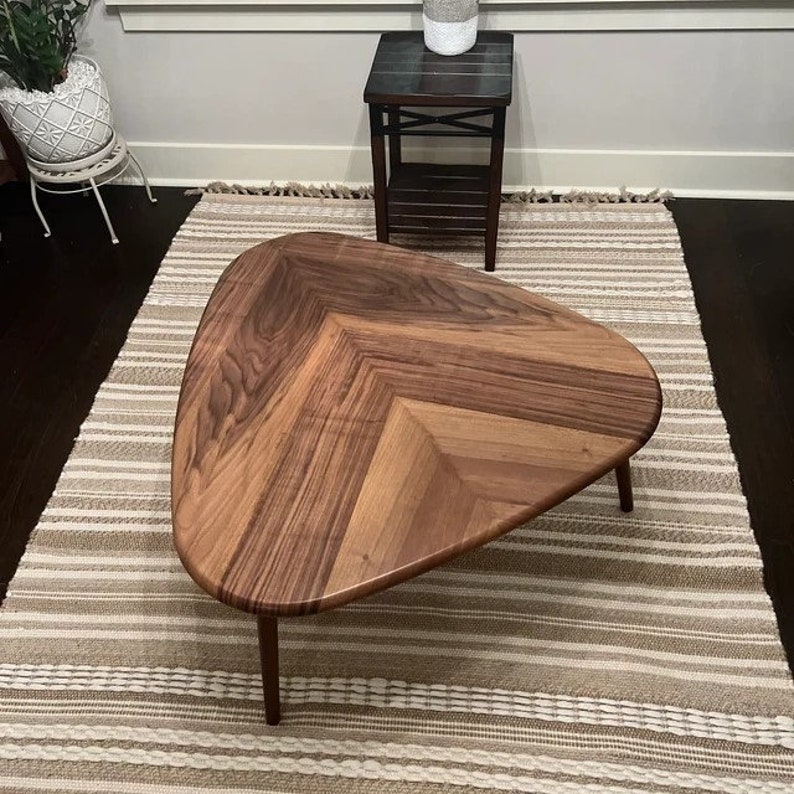 May include: Triangular wooden coffee table with a chevron pattern. The table has a natural wood finish and sits on four slender legs. The table is placed on a striped rug.