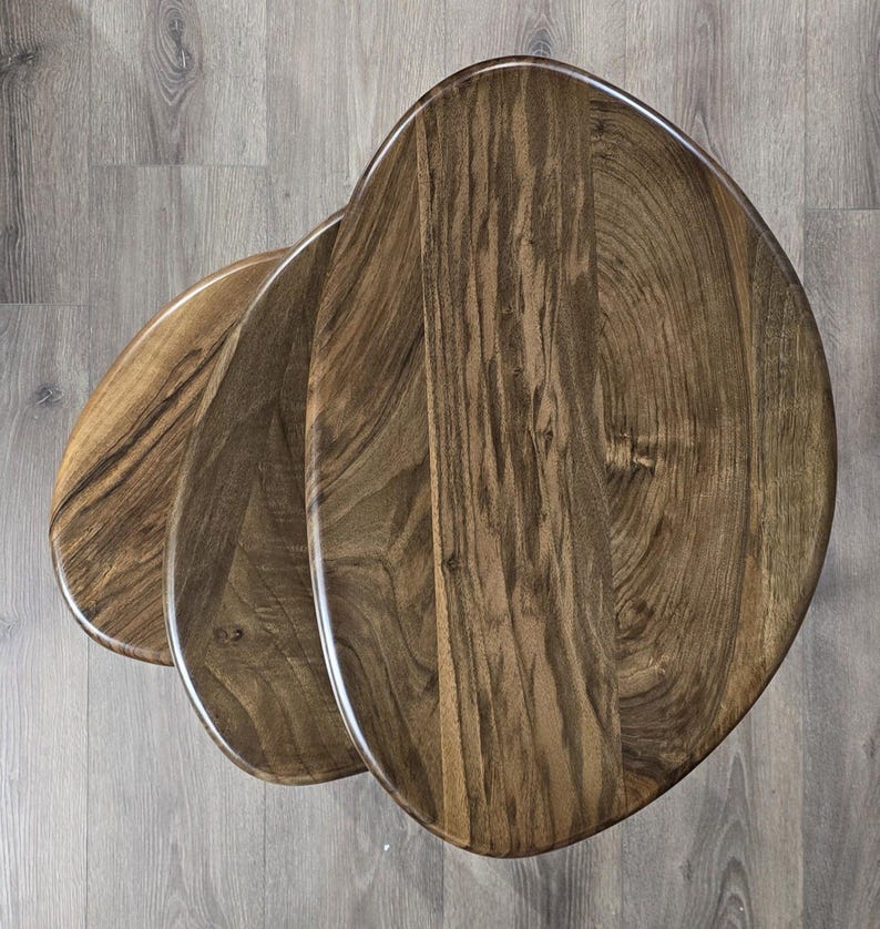 May include: A set of three wooden nesting tables with a unique organic shape. The tables are made of dark brown wood with a natural grain pattern.