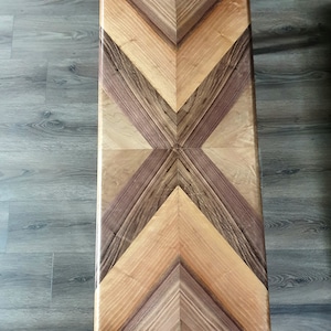 May include: Rectangular wooden coffee table with a geometric pattern. The tabletop features a symmetrical design of light and dark wood in shades of brown and tan. The table's surface is smooth and polished, showcasing the natural wood grain.
