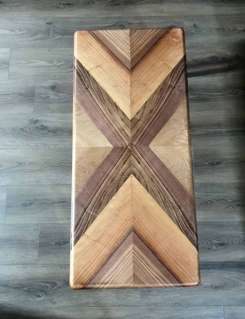 May include: Rectangular wooden coffee table with a geometric pattern. The tabletop features a symmetrical design of light and dark wood in shades of brown and tan. The table's surface is smooth and polished, showcasing the natural wood grain.