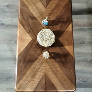 May include: A rectangular wooden coffee table with a chevron pattern in shades of brown. Three decorative items are placed on the table: a blue and silver pendant, a woven coaster, and a gold sphere.