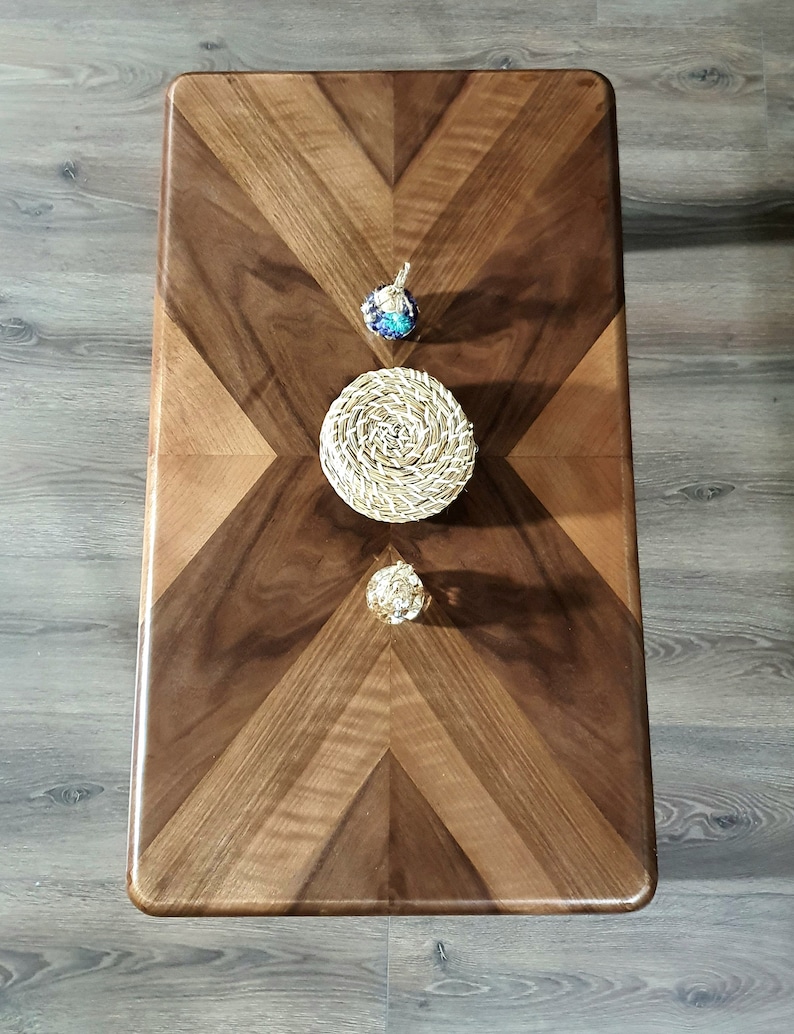 May include: A rectangular wooden coffee table with a chevron pattern in shades of brown. Three decorative items are placed on the table: a blue and silver pendant, a woven coaster, and a gold sphere.