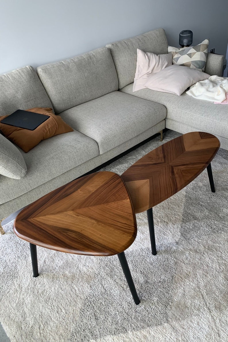 May include: Two wooden coffee tables with a geometric design and black legs. The tables are different shapes, one is triangular and the other is oval. The tables are placed on a light gray rug in front of a gray sofa.