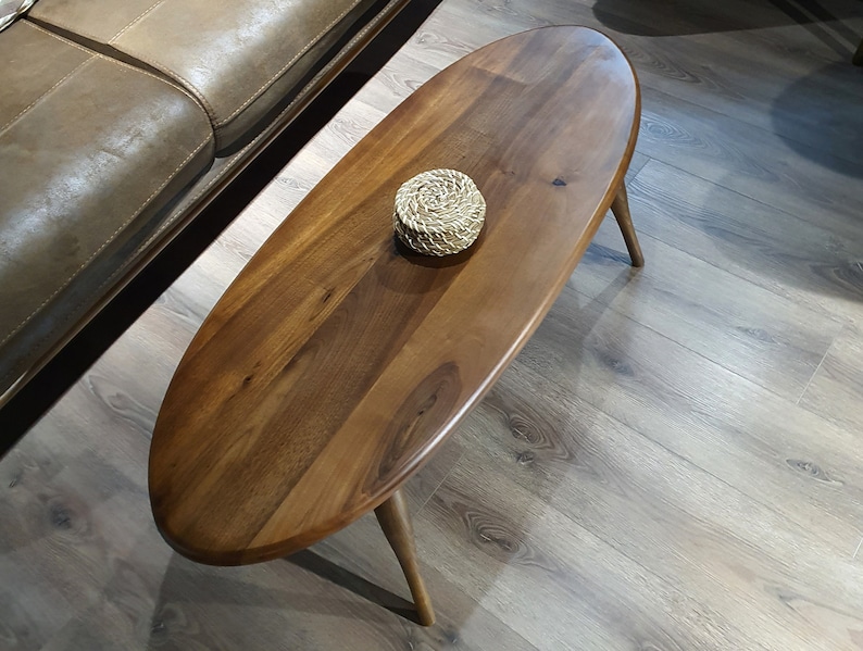 May include: A dark brown oval-shaped wooden coffee table with a woven coaster on top. The table has four legs and is sitting on a light brown wood floor.