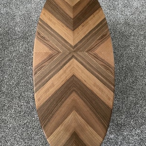 May include: Oval wooden coffee table with a chevron pattern. The tabletop features alternating bands of light and dark wood, creating a striking visual design. The table's shape is elongated, with rounded ends.