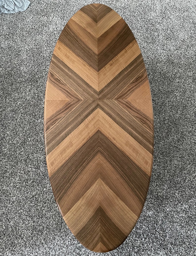 May include: Oval wooden coffee table with a chevron pattern. The tabletop features alternating bands of light and dark wood, creating a striking visual design. The table's shape is elongated, with rounded ends.