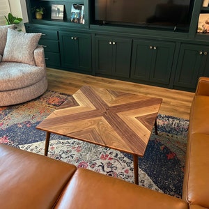 May include: A square wooden coffee table with a geometric pattern of light and dark wood. The table has a modern design and is sitting on a rug with a colorful pattern.